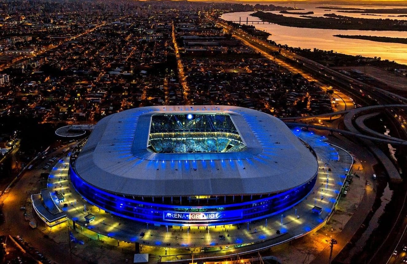 Arena do Grêmio lit in blue at night, seen from the air with the city and the river behind