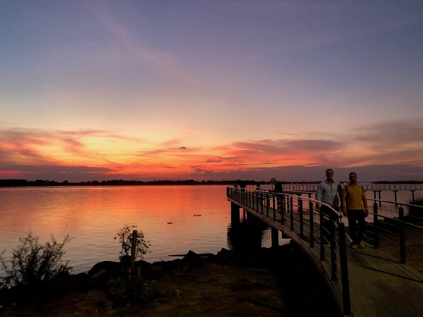 Sunset over the Guaíba seen from a waterfront deck, with people watching from the railing